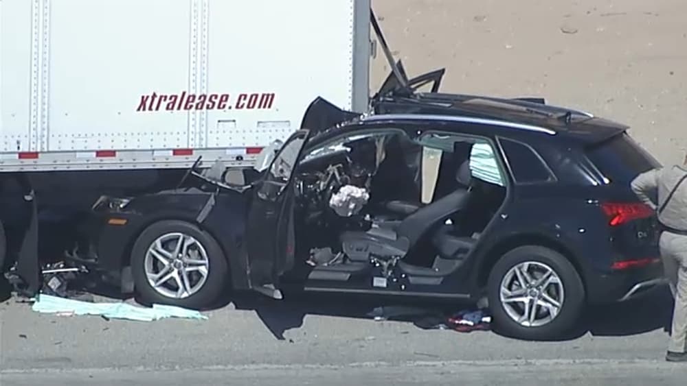 A black Audi SUV with its entire front end sheared away, resting beside the rear of a white semi-trailer on a freeway.