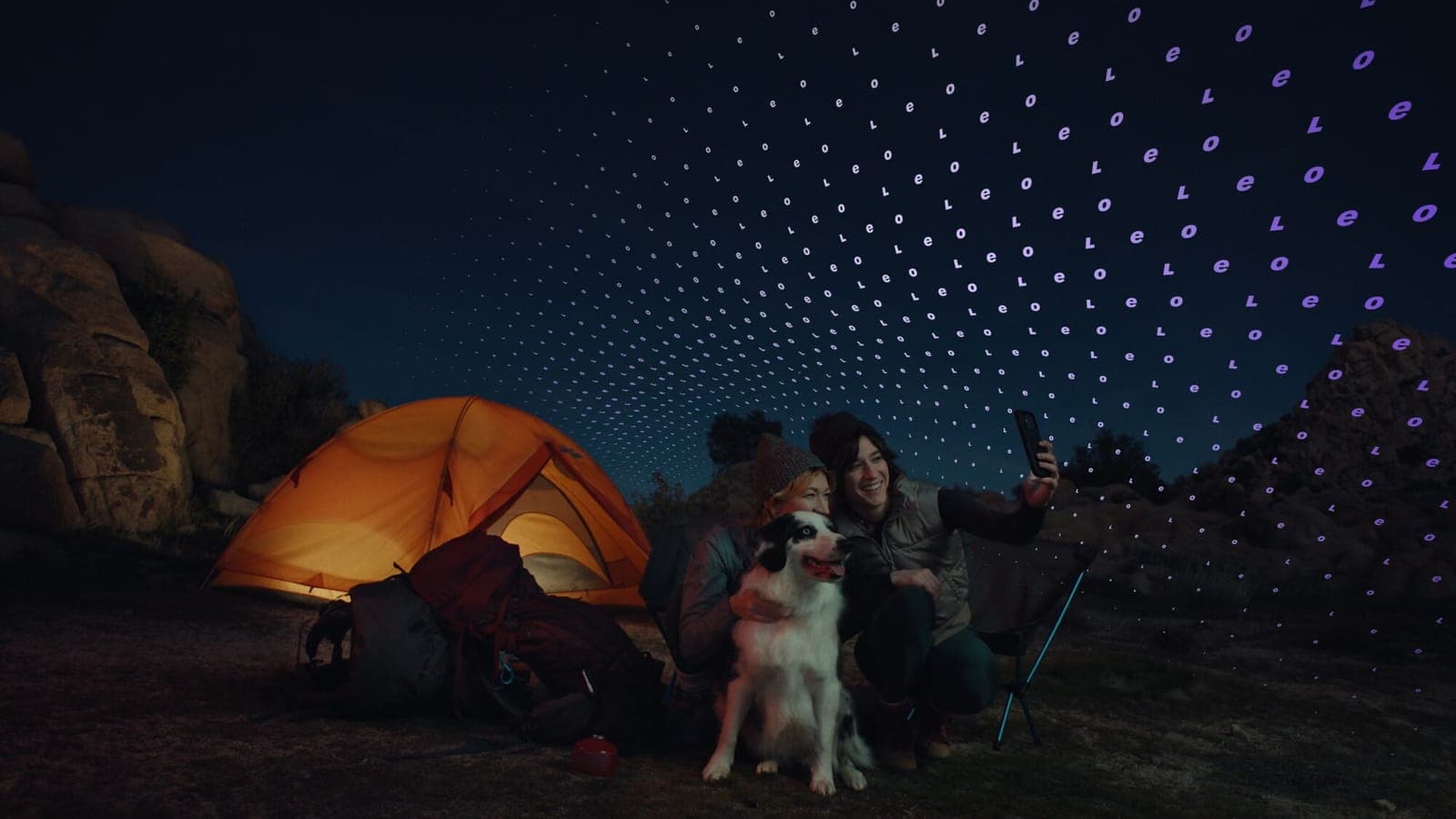 Two people and a husky sitting beside an illuminated orange tent at night under a star-filled sky.