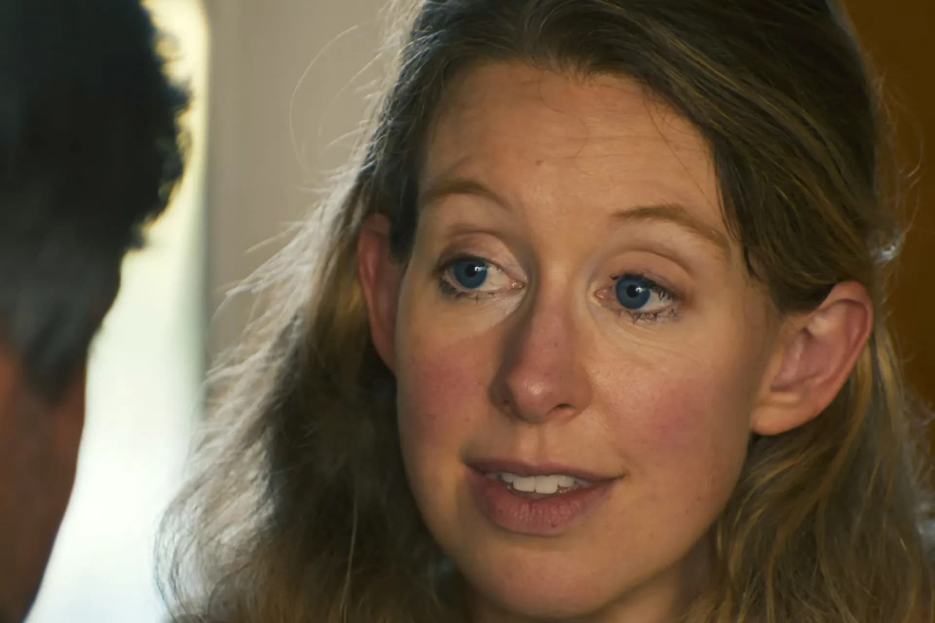 Elizabeth Holmes, with light hair, looking toward someone off camera in a still from the documentary.