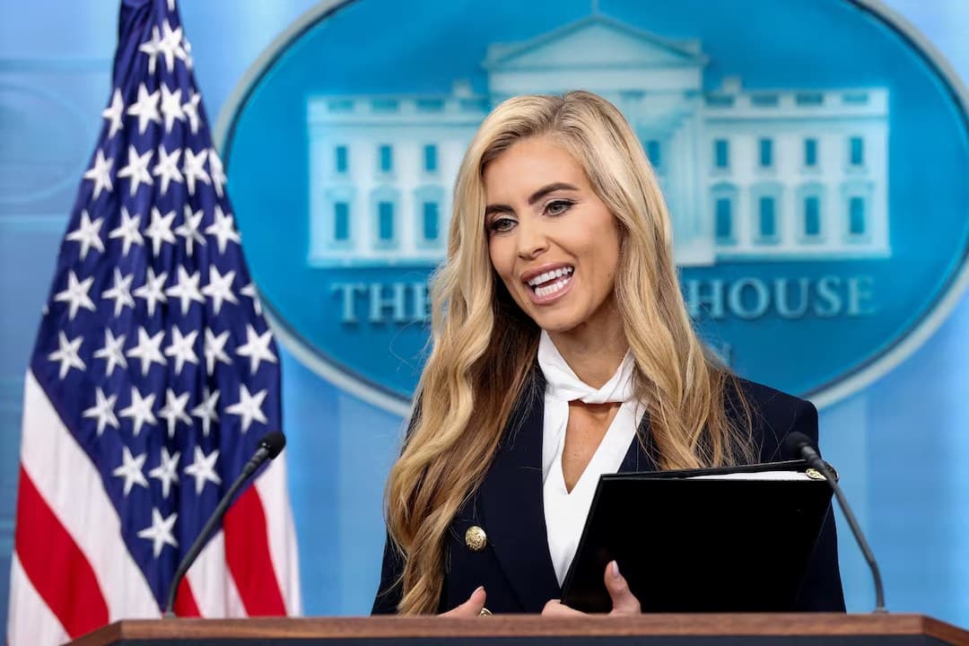 Katie Zacharia, in a dark jacket with gold buttons, stands at the White House briefing room podium in front of an American flag.