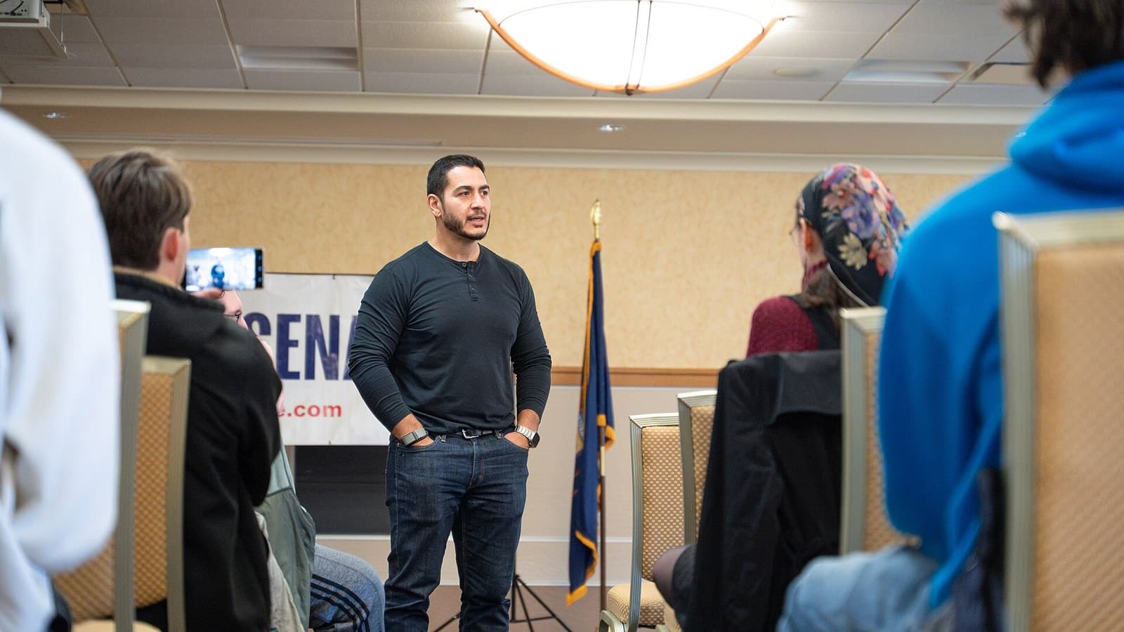 Abdul El-Sayed, in a dark long-sleeved shirt and jeans, stands with his hands in his pockets speaking to a seated audience in a conference room, with a Michigan flag behind him.