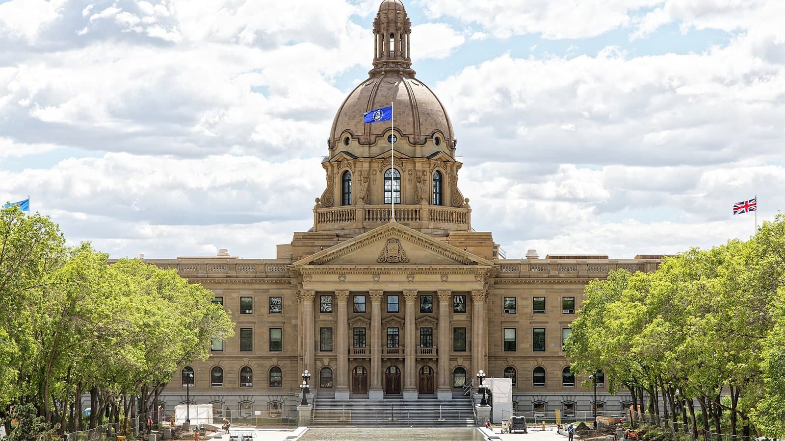 The domed sandstone Alberta Legislature Building in Edmonton under a partly cloudy sky, with trees lining the plaza in front.