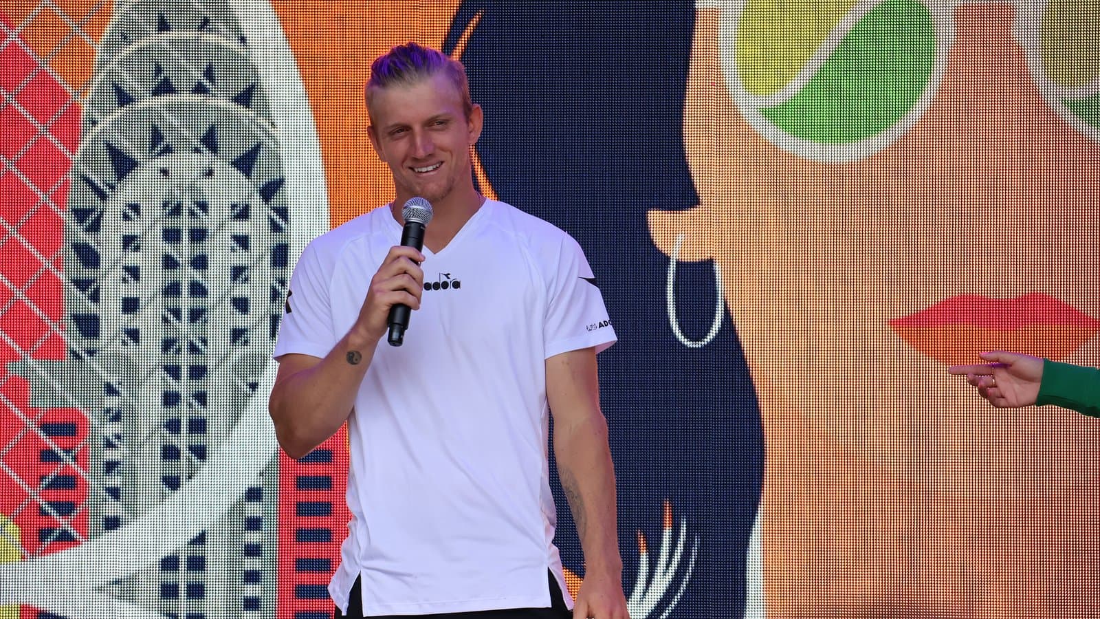 Spanish tennis player Alejandro Davidovich Fokina in a white T-shirt smiling and speaking into a microphone in front of a colorful mural.