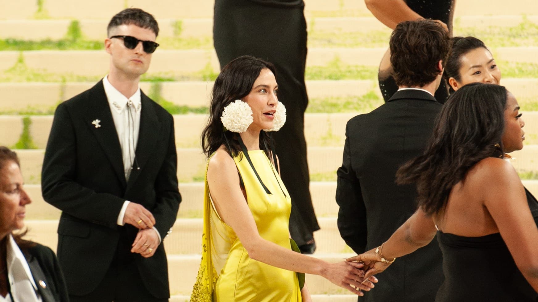 Alexa Chung in a long yellow satin gown with white flower ear decorations, smiling and reaching to take another guest's hand on the Met Gala steps, with guests in black around her.