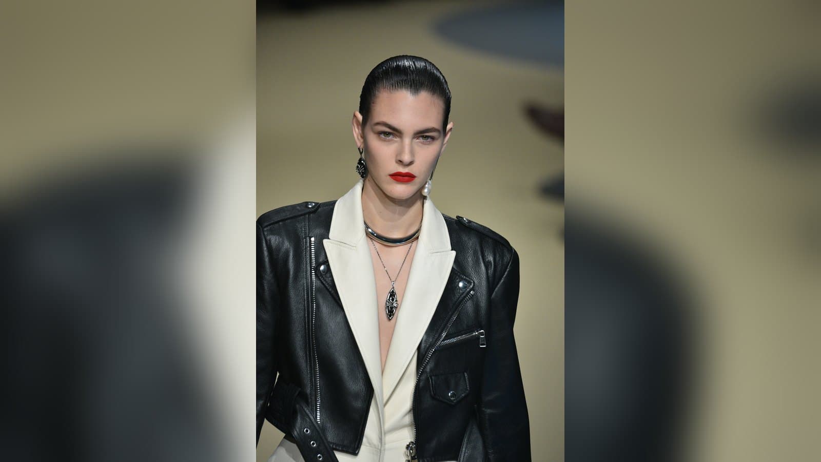 A model with slicked-back hair and red lipstick walks a runway in a black leather biker jacket over a cream tailored jacket and layered necklaces.