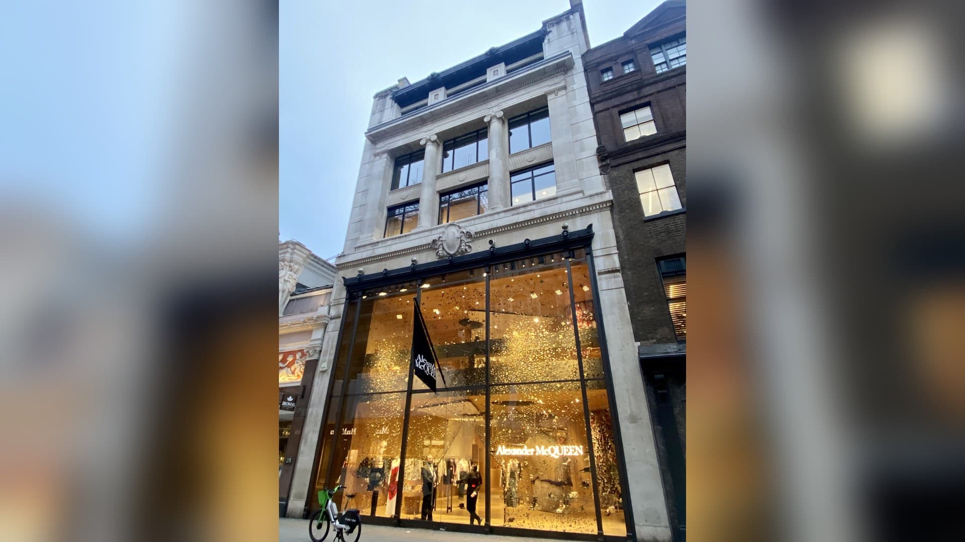 The Alexander McQueen store on Old Bond Street in London: a pale stone building with a double-height glass shopfront, a black McQueen flag and a window display of falling gold petals.