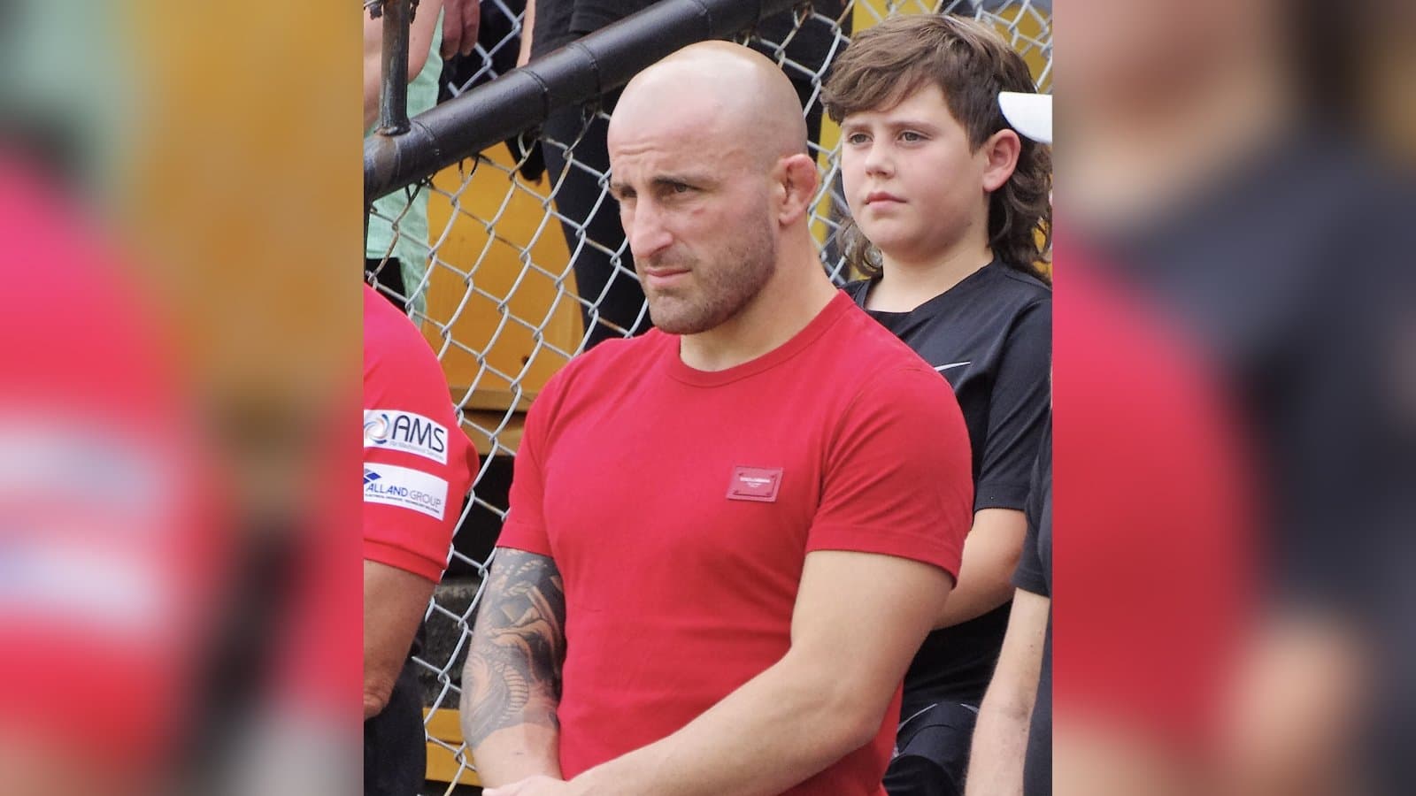 UFC featherweight champion Alexander Volkanovski in a red T-shirt standing beside the cage fence.