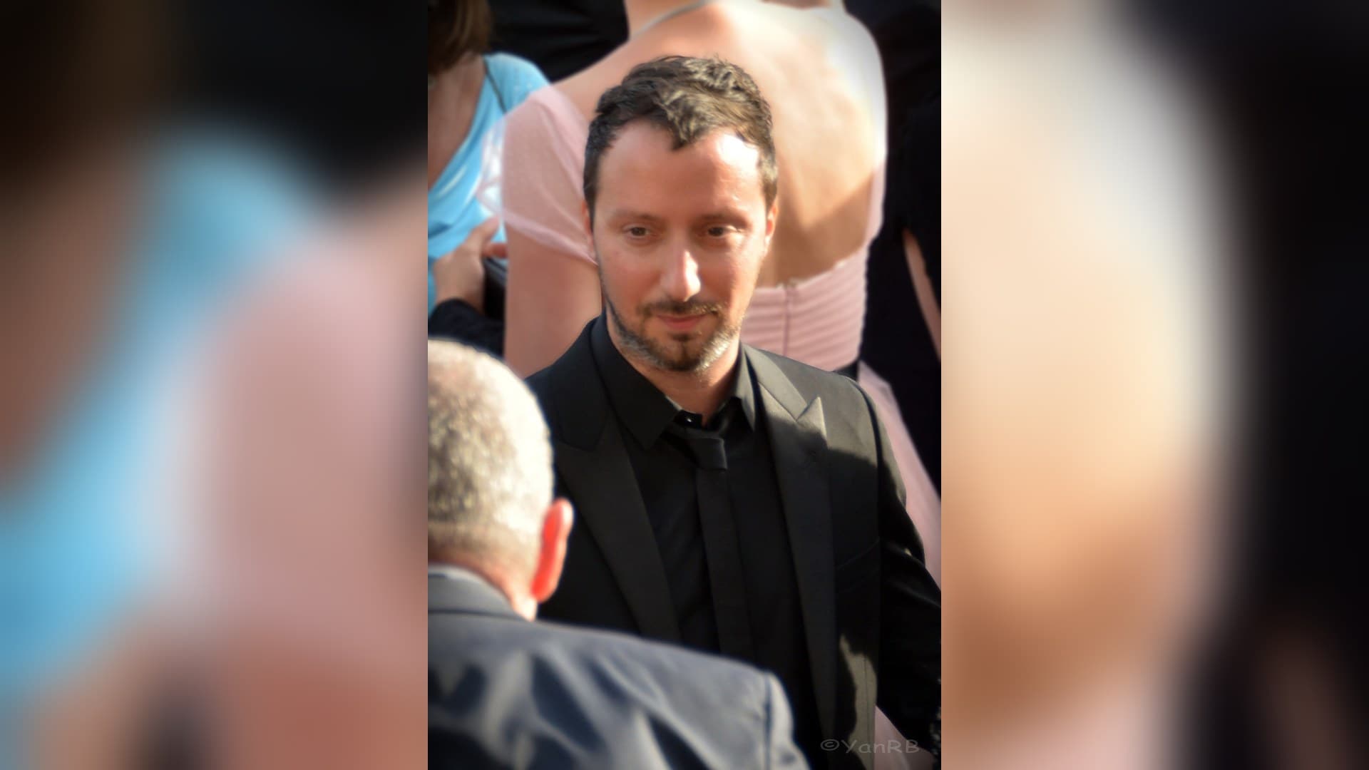 Designer Anthony Vaccarello, with short dark hair and a beard, wearing a black suit, black shirt and black tie on the red carpet at Cannes.