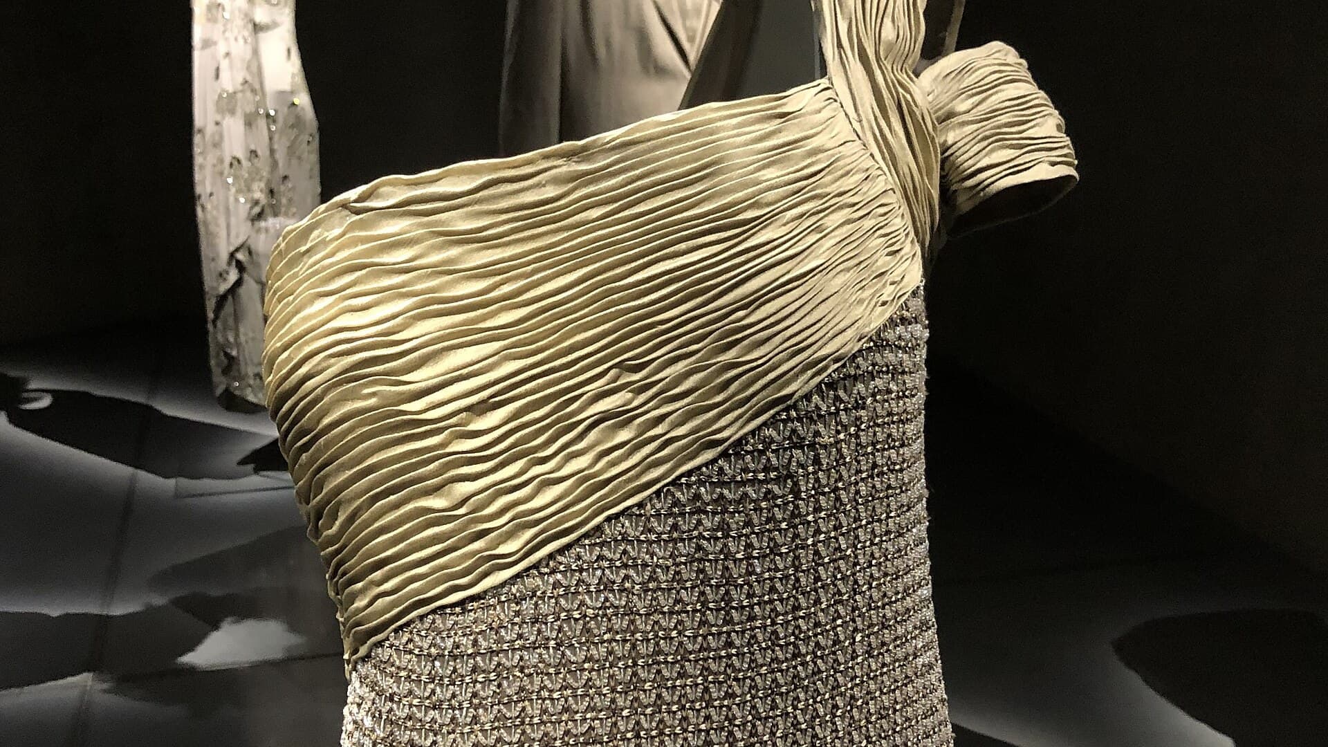 Close-up of a sand-coloured pleated one-shoulder gown with a beaded lower section, displayed on a mannequin against a dark background at the Armani/Silos museum.