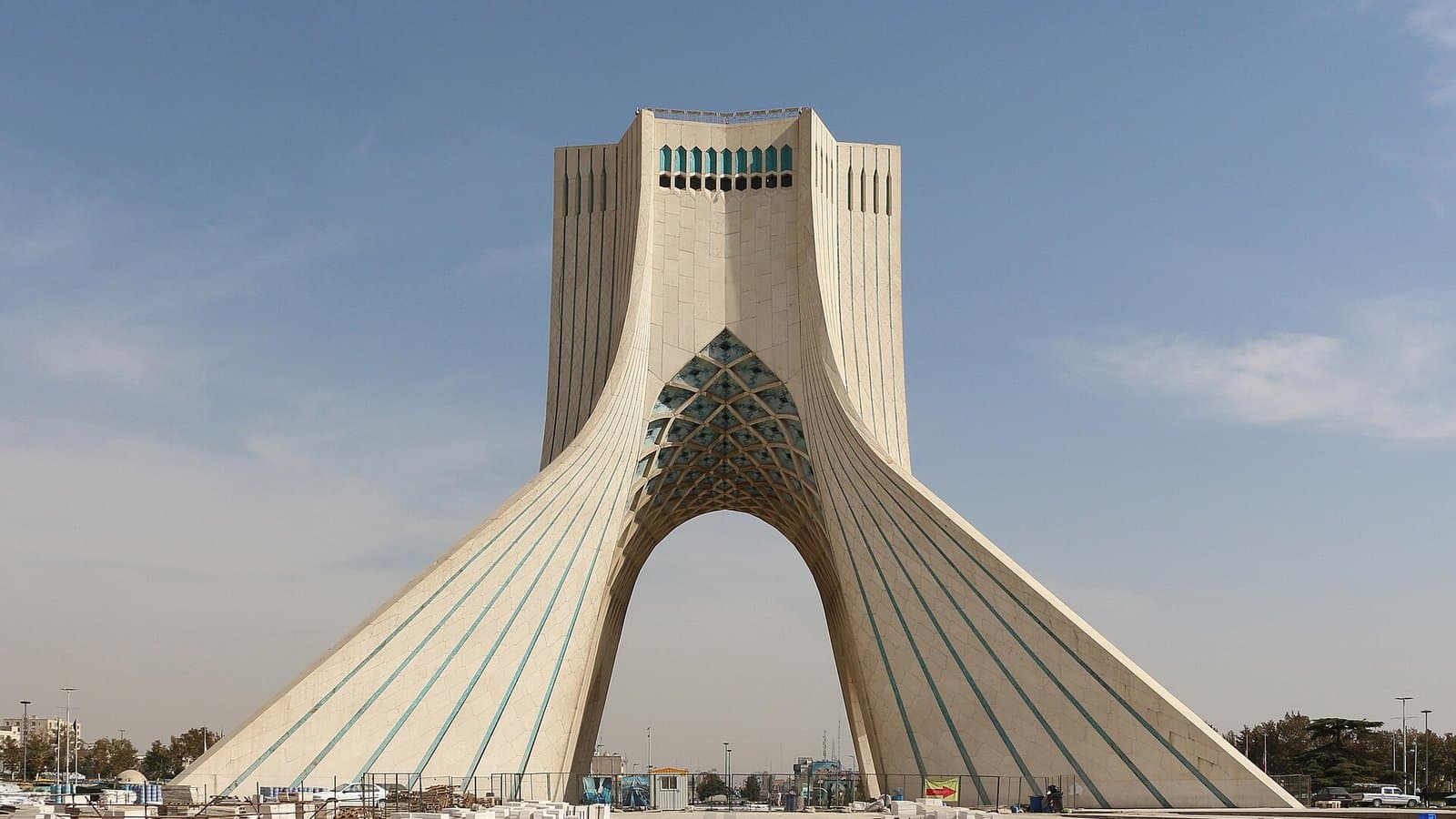 Tehran's white marble Azadi Tower, an arched monument with a wide base and tapering top, under a blue sky with light clouds.