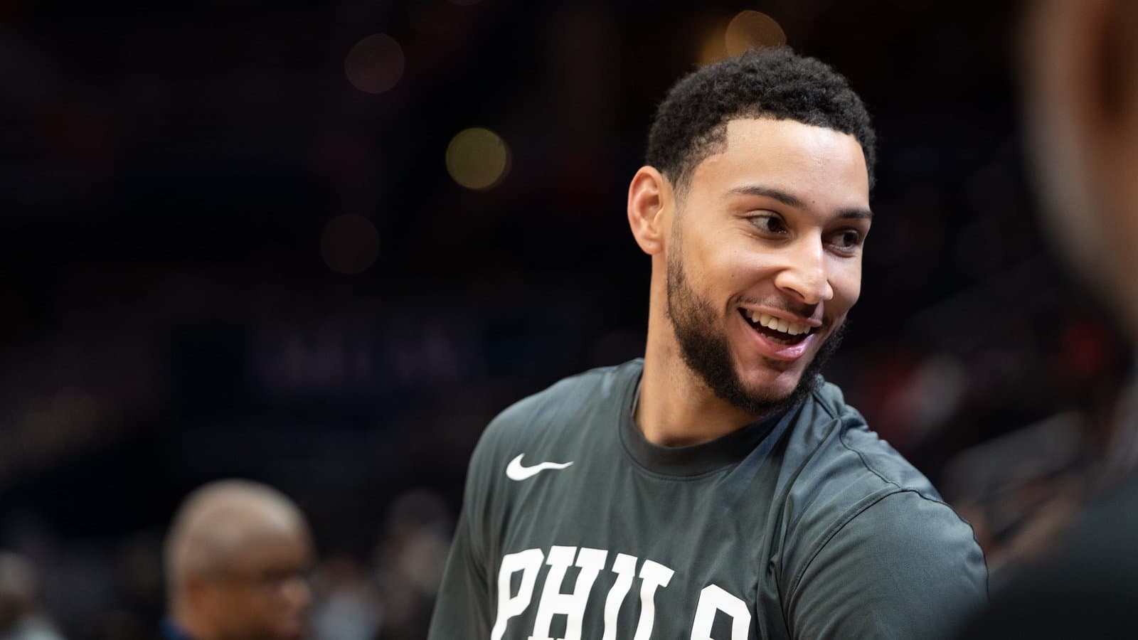Ben Simmons smiling in a dark Philadelphia warm-up shirt on the court before a game.