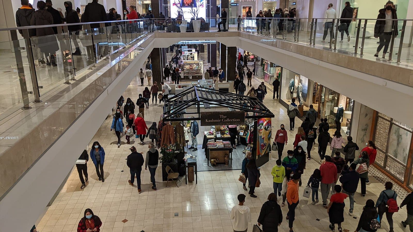 View from an upper floor of a busy two-level shopping mall on Black Friday, with crowds of shoppers walking past kiosks below and store fronts and railings on both sides.