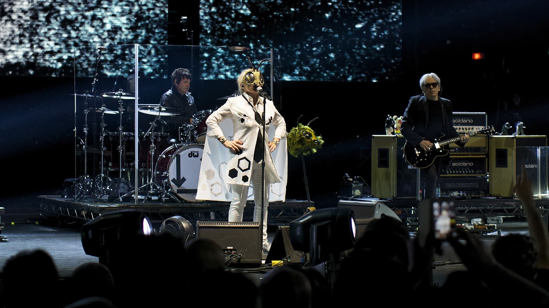 Debbie Harry in a white coat and a gold bee mask sings at a microphone, with a drummer behind her and Chris Stein playing a black guitar on the right, in front of a dark crowd.