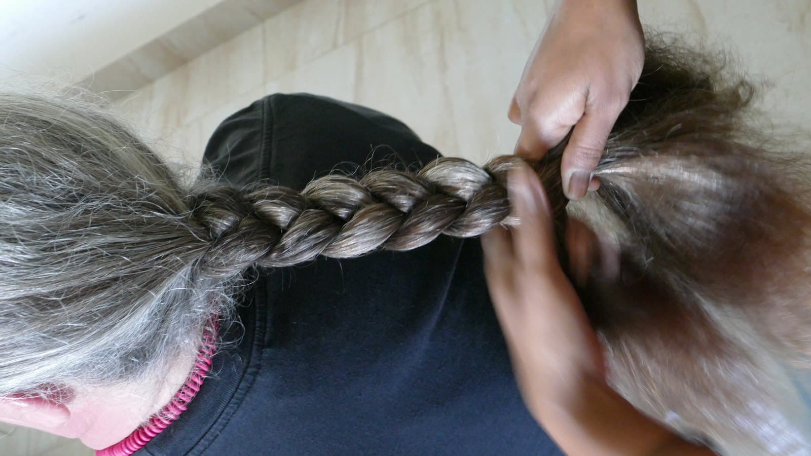 Close-up of hands weaving a long brown-and-grey plait at the back of a person's head.