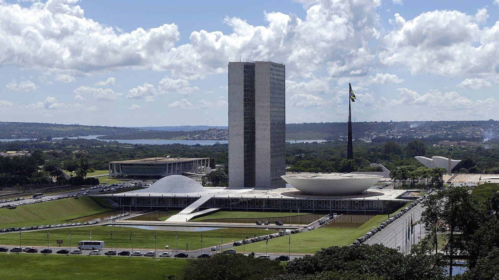 Brazil's National Congress complex in Brasília, with its twin office towers and two bowl-shaped chambers on a wide green lawn under a blue sky.