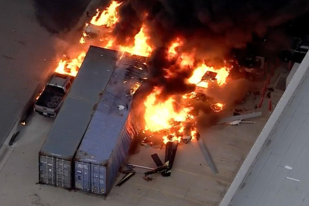 Aerial view of a fiercely burning aircraft wreck beside two shipping containers and a burning pickup truck in a parking lot, with debris scattered across the asphalt.