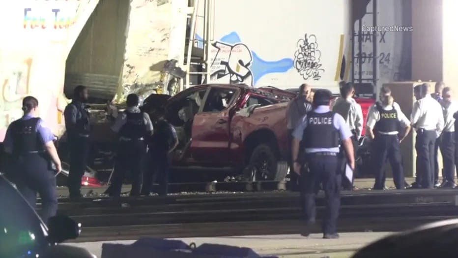 Emergency responders gathered around a wrecked vehicle beside a freight rail car.