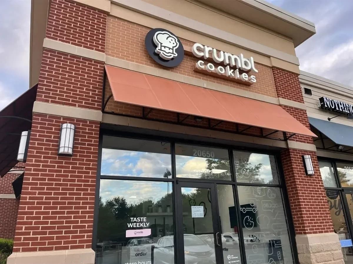 A brick storefront with a coral awning and a Crumbl Cookies sign with the chain's chef-hat logo above glass doors.