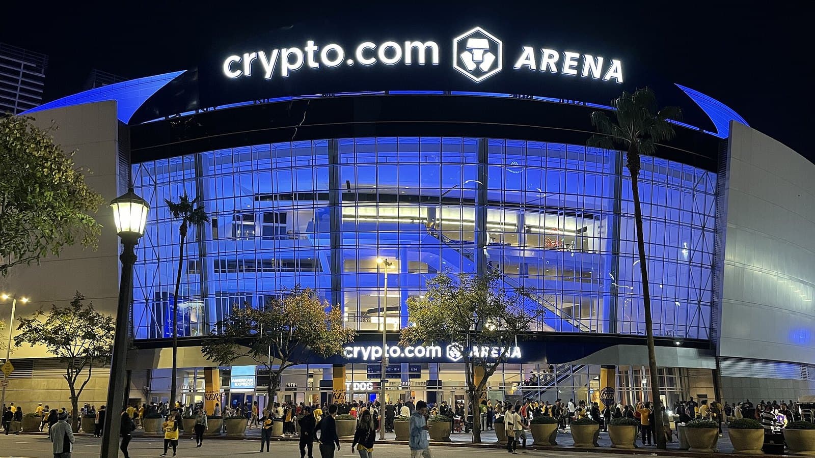 The glass front of Crypto.com Arena lit in blue at night, with its illuminated name across the roof and crowds and palm trees on the plaza outside.