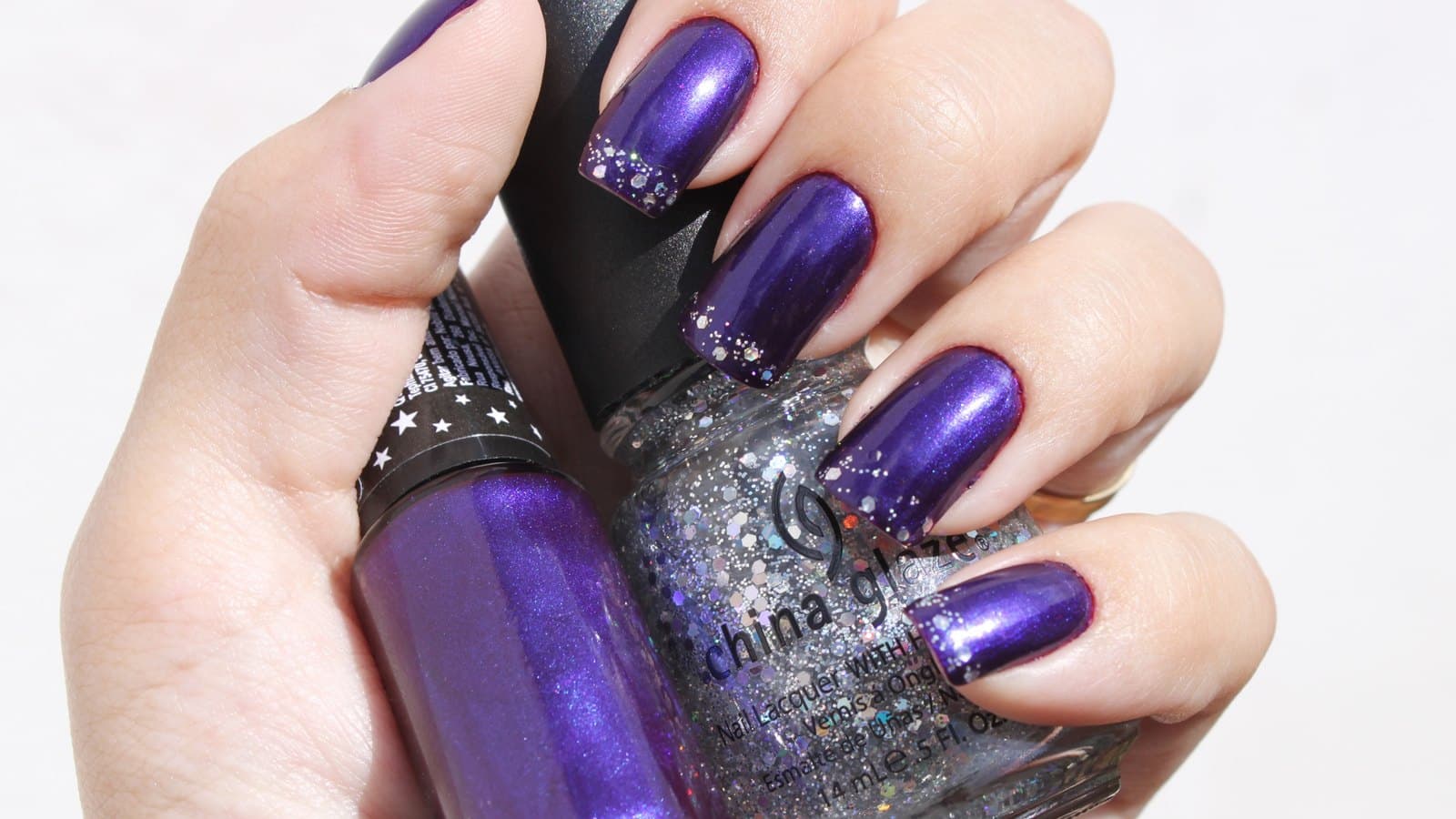 A hand with glossy deep violet nails and silver glitter tips holding two bottles of purple and glitter nail polish against a white background.