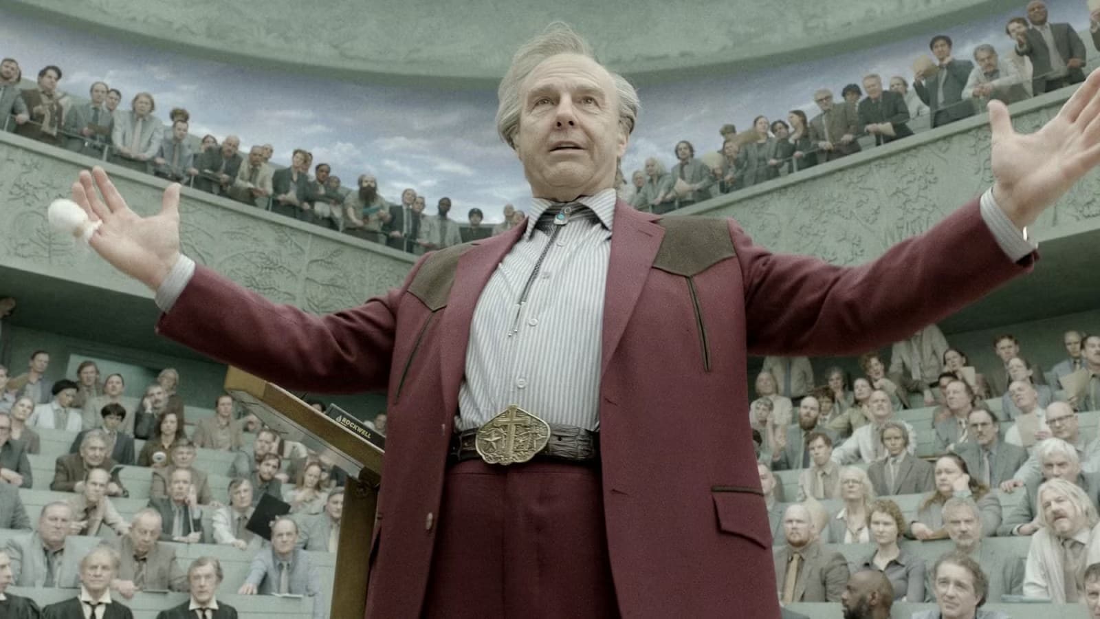 Tom Cruise, with gray hair and a burgundy suit, stands with his arms spread wide in front of a crowd in a stadium.