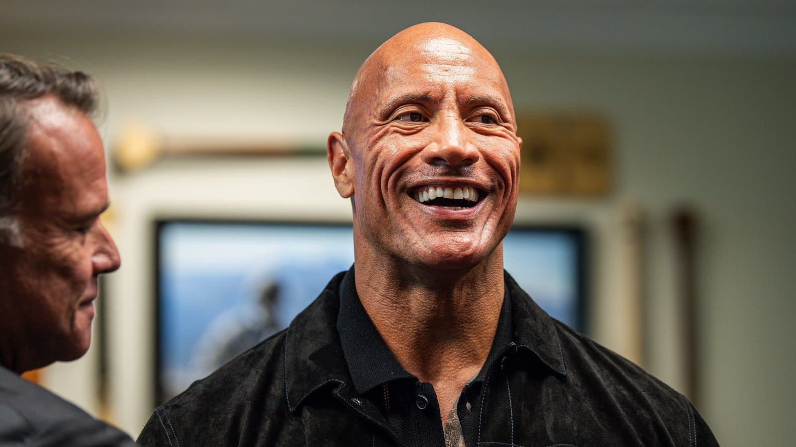 Dwayne Johnson, bald and smiling broadly, wearing a black suede jacket, talks with a man indoors.