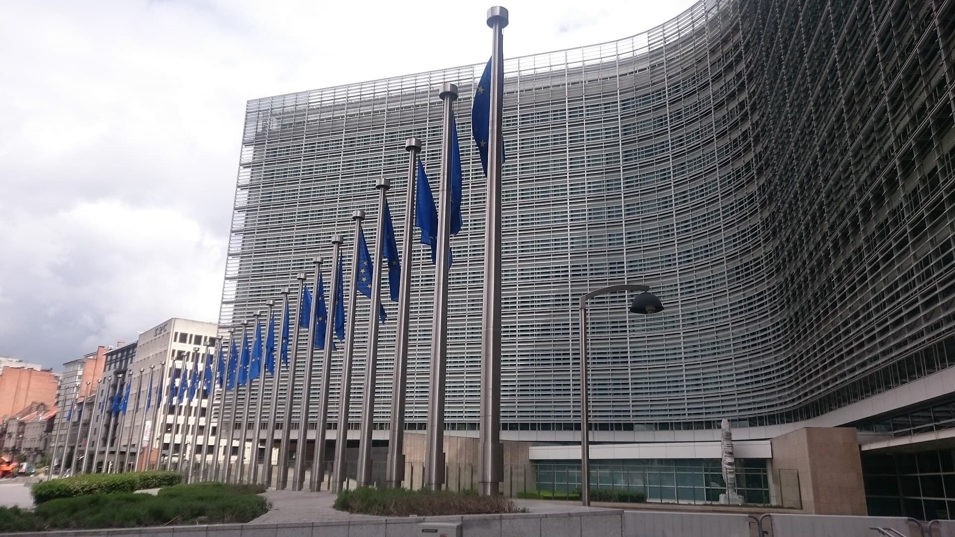 A row of blue European Union flags on tall poles in front of the curved glass facade of the Berlaymont building in Brussels under a cloudy sky.