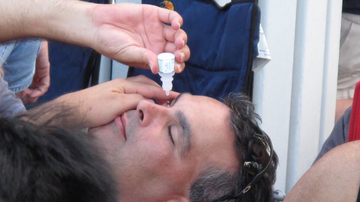 A man tilting his head back and squeezing a drop from a small plastic bottle into his eye.