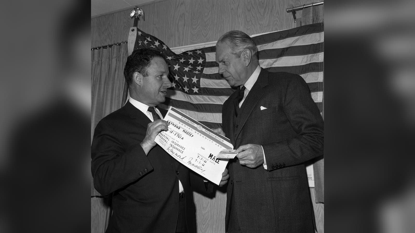 Black-and-white photo of two men in suits in front of an American flag, one holding up an oversized Medicare identification card while the other, older man looks at it.