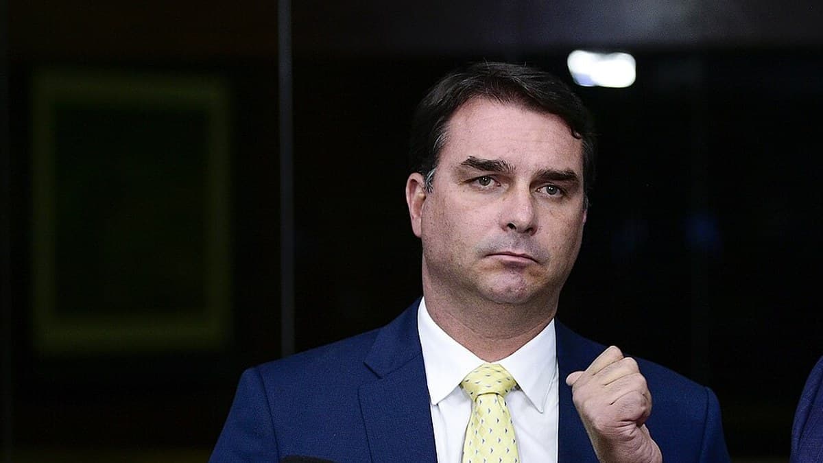 Flávio Bolsonaro, in a navy suit and pale yellow tie, raising a clenched fist as he speaks behind a cluster of broadcast microphones.