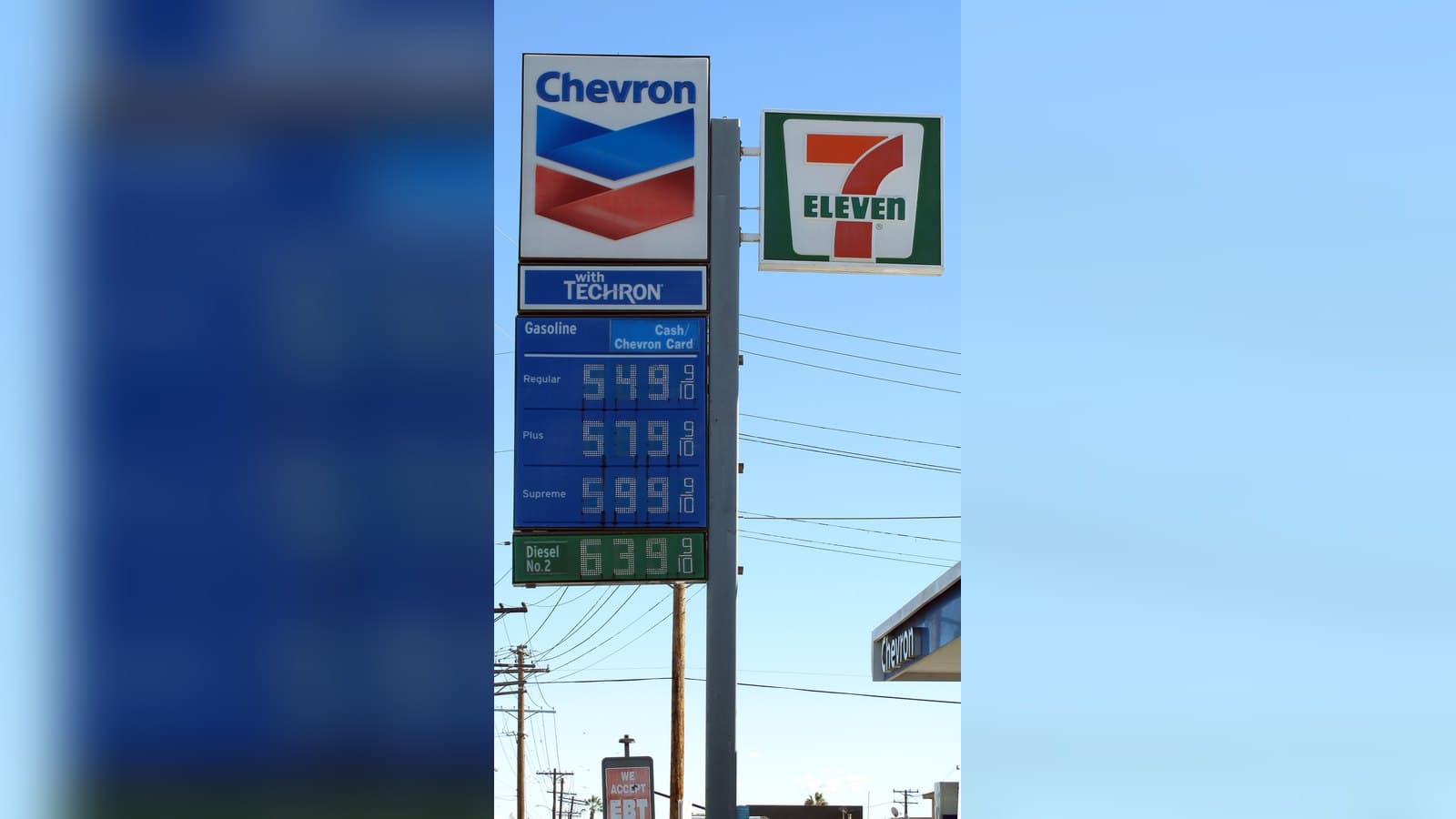 A tall Chevron gas station sign against a blue sky listing gasoline prices of $5.49, $5.79 and $5.99 a gallon and diesel at $6.39, next to a 7-Eleven sign.