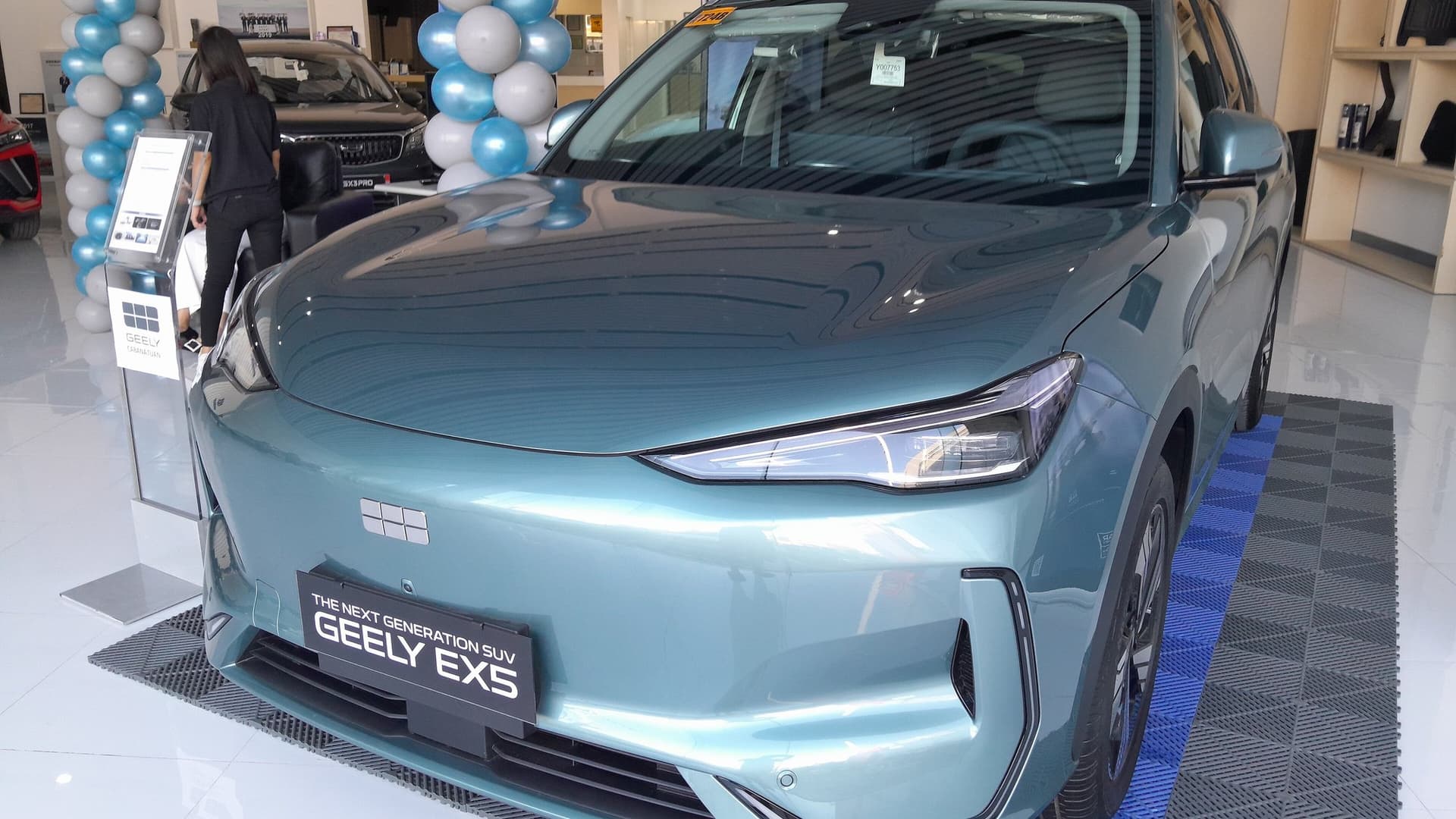 A teal Geely EX5 electric SUV on a dealership showroom floor, with a sign reading The Next Generation SUV Geely EX5 and blue and white balloons behind it.