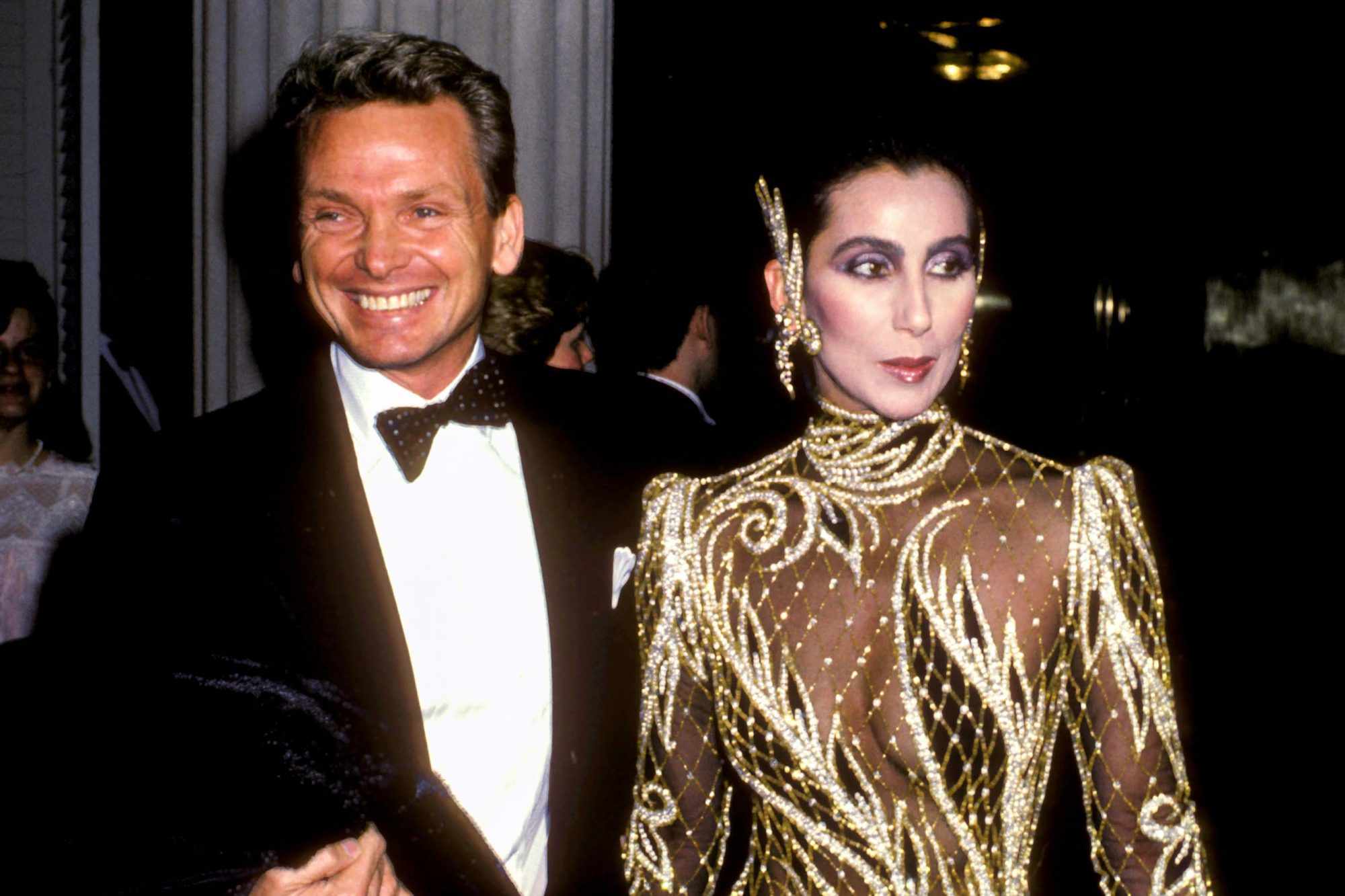 Bob Mackie in a tuxedo, smiling, beside Cher in a sheer gold-and-pearl beaded high-necked gown.