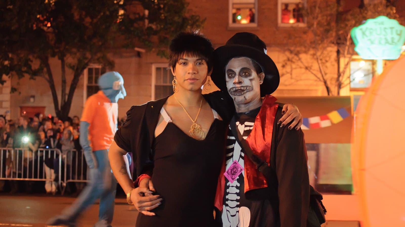 Two people in Halloween costumes, one with skeleton face paint and a top hat, pose arm in arm on a New York street during the Village Halloween Parade at night.