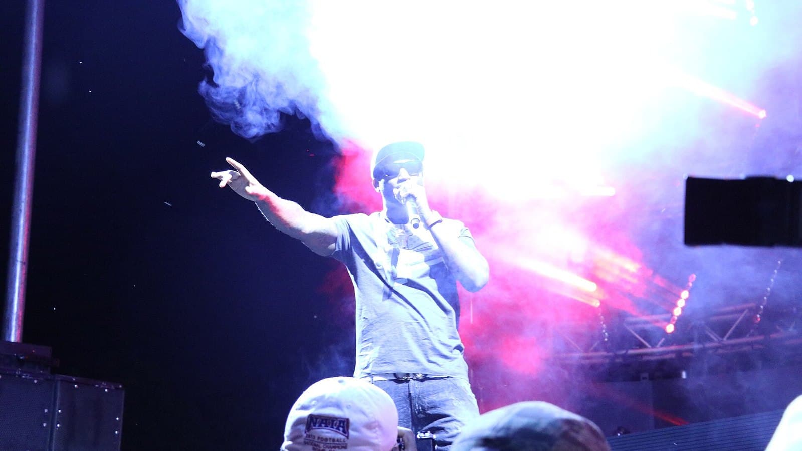 Gucci Mane in a cap and grey T-shirt points toward the crowd with a microphone in hand as pink and white smoke billows across an outdoor festival stage at night.