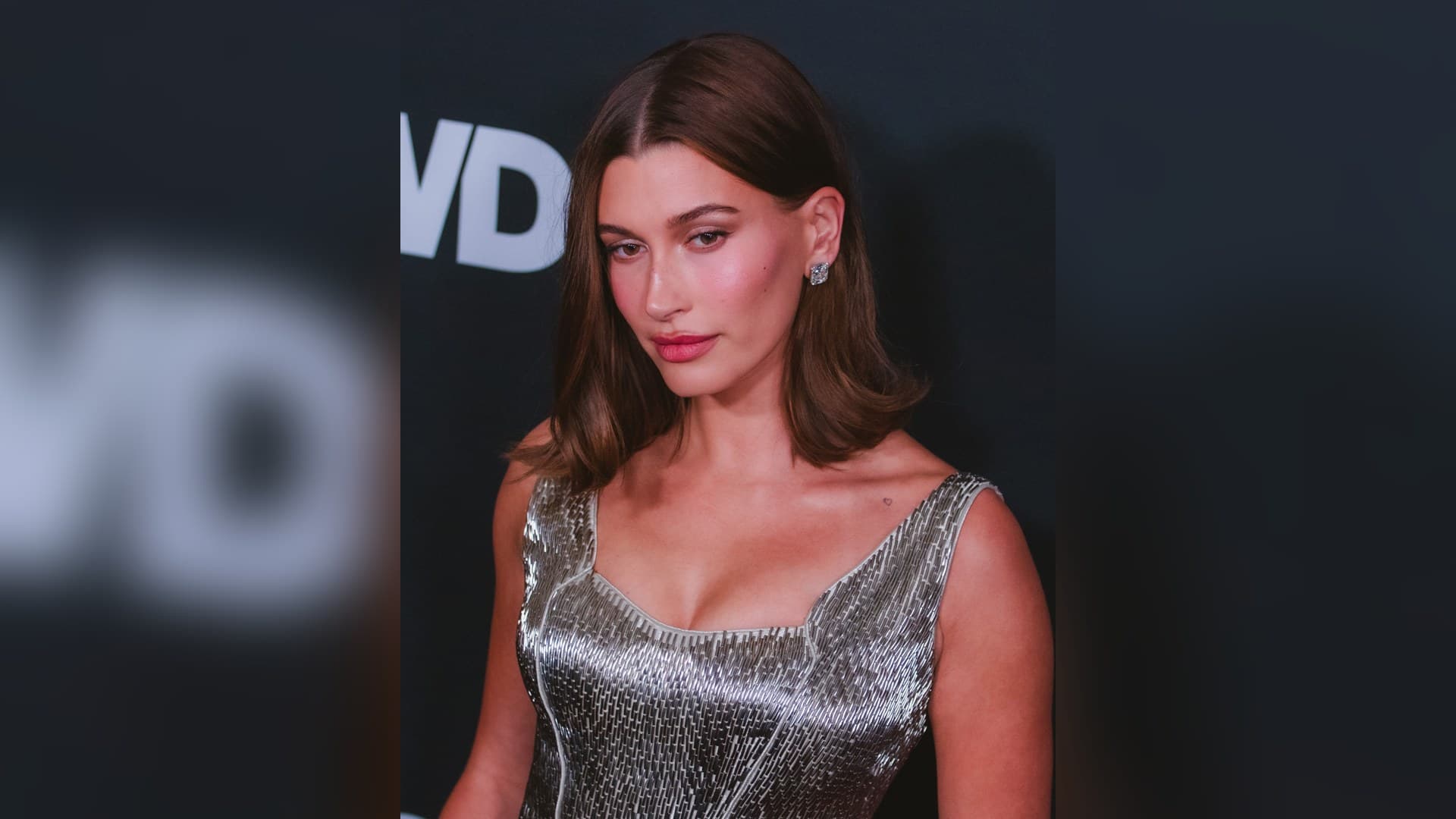 Hailey Bieber with shoulder-length brown hair and a sparkling diamond earring, wearing a beaded silver sleeveless dress in front of a dark WWD backdrop.