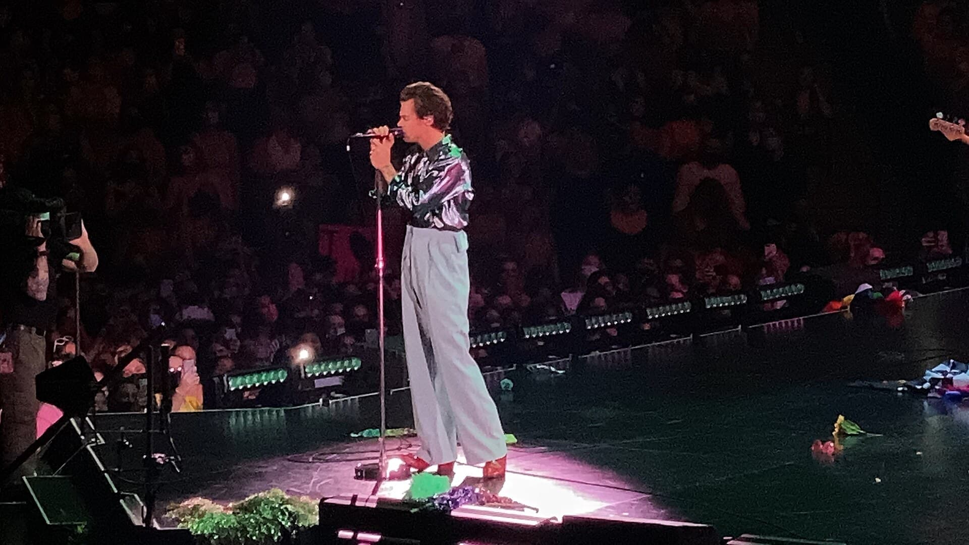 Harry Styles in a shimmering shirt and wide pale trousers sings into a microphone on a stand, lit by a pink spotlight in front of a packed arena crowd.