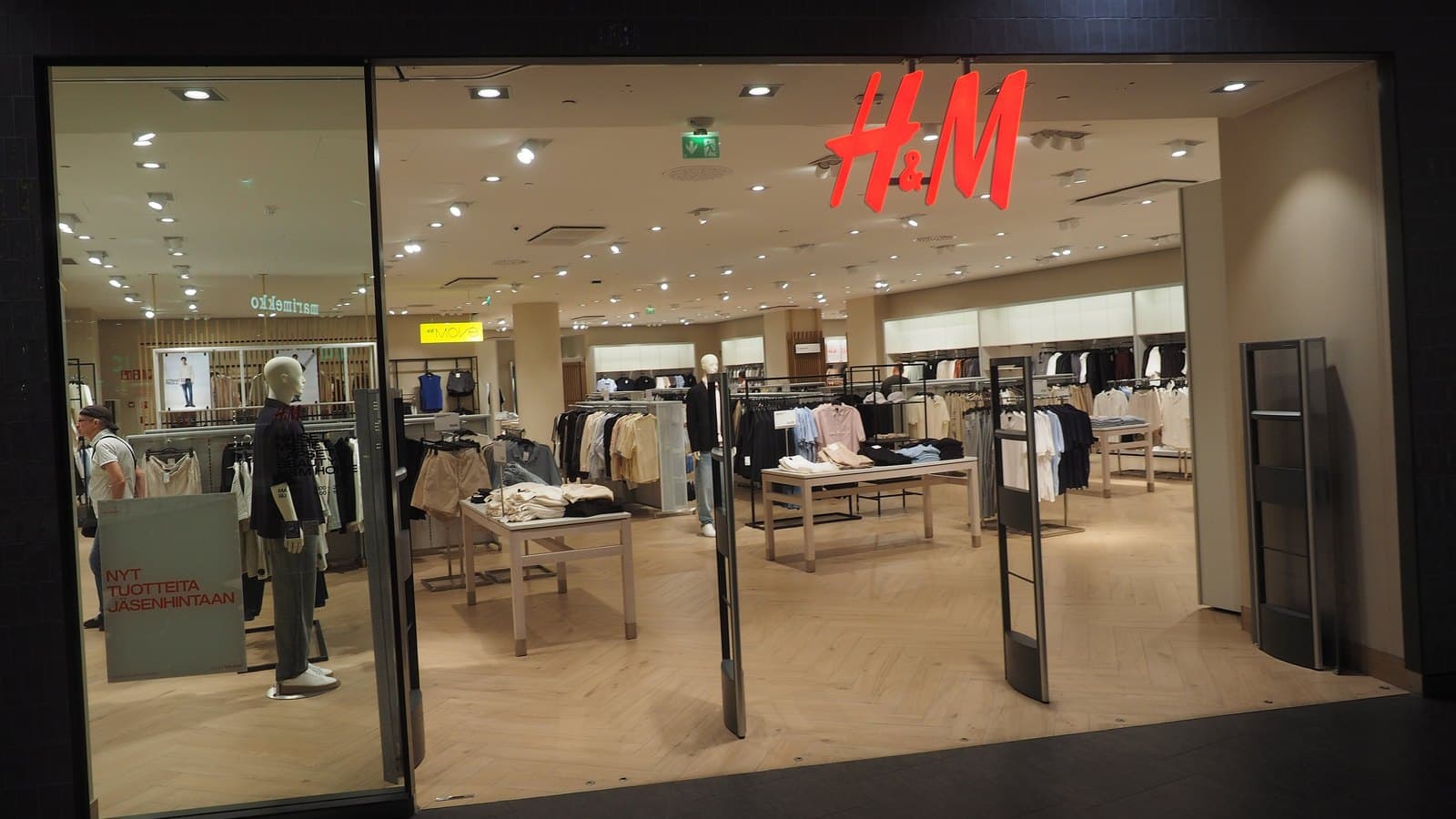 The glass entrance of an H&M store inside a shopping centre, with the red H&M logo above and racks of clothing and mannequins visible inside.