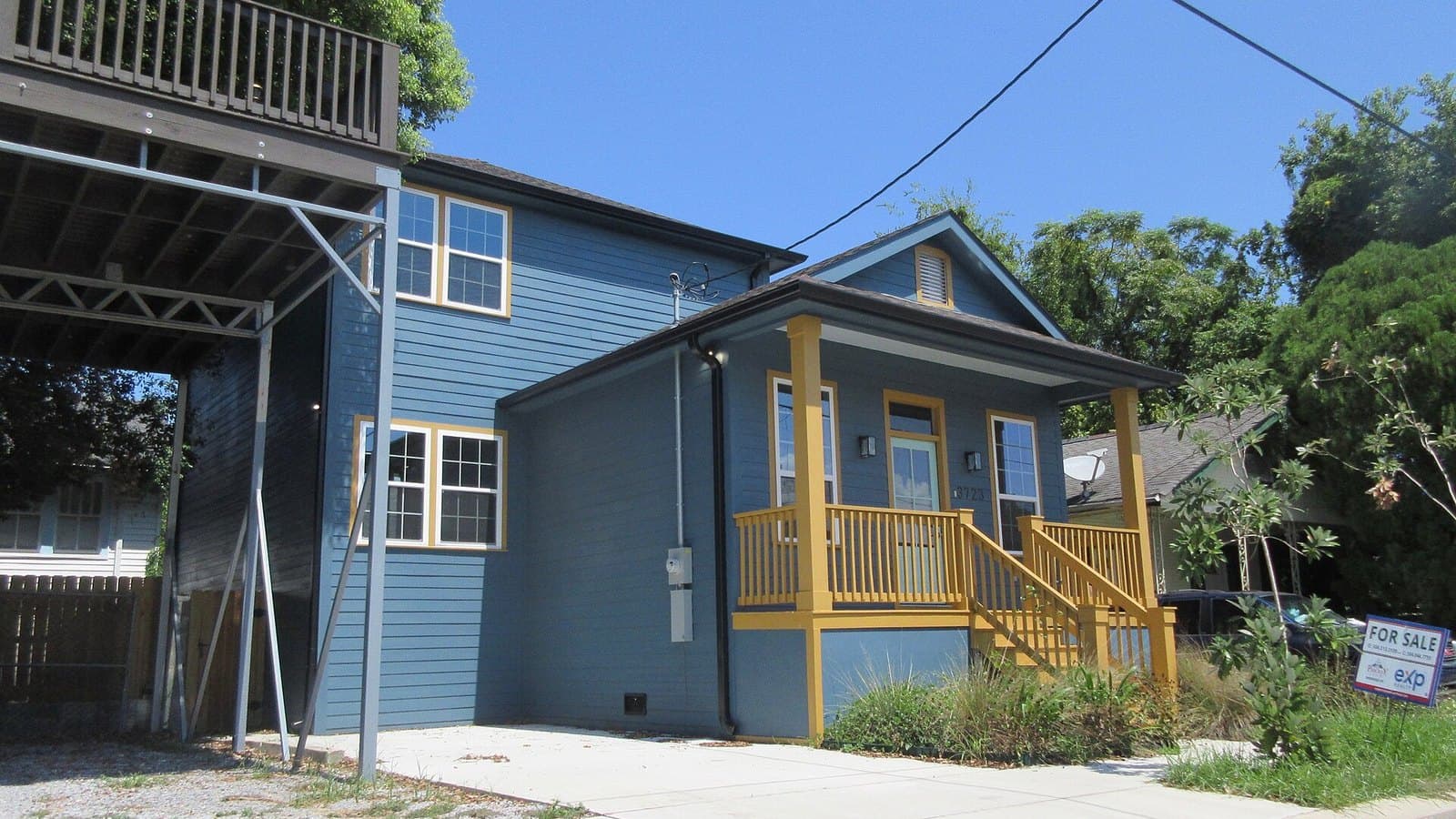 A new two-storey blue house with a yellow-trimmed front porch and steps, a small for-sale sign in the front yard and trees behind it under a clear blue sky.