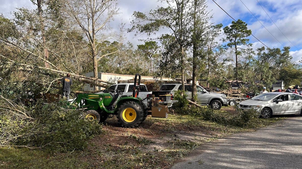 Storm damage in East Milton, Florida, after Hurricane Isaias, with broken trees and debris around buildings under a grey sky.