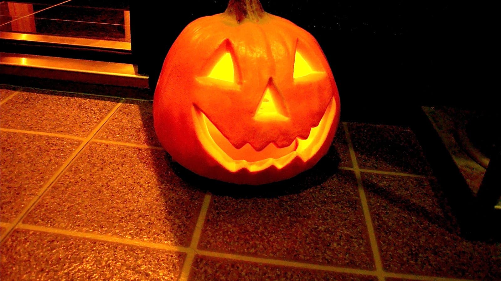 A glowing carved pumpkin with triangle eyes and a toothy smile sitting on a tiled step outside a doorway at night.