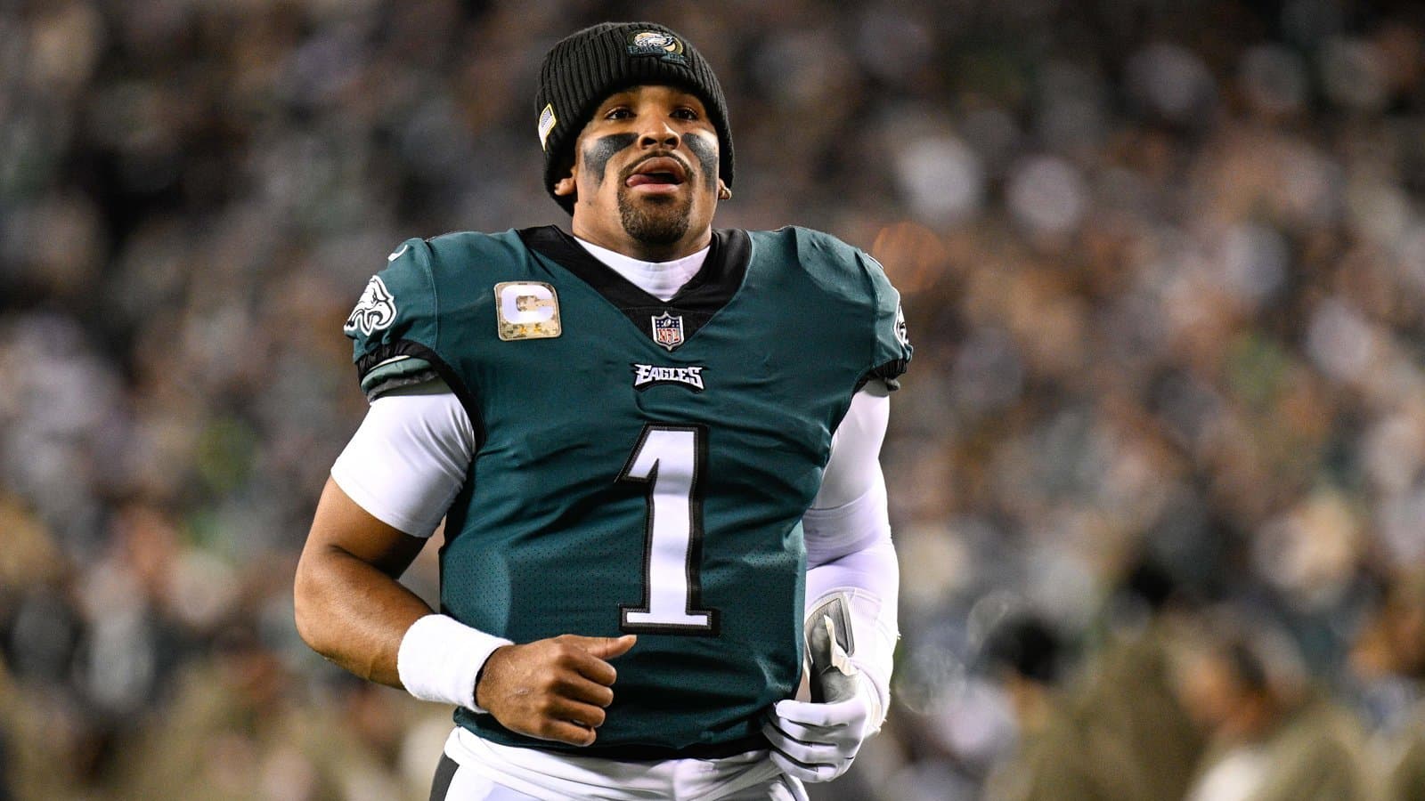 Philadelphia Eagles quarterback Jalen Hurts in a green No. 1 jersey and black beanie jogging on the field in front of a crowd.