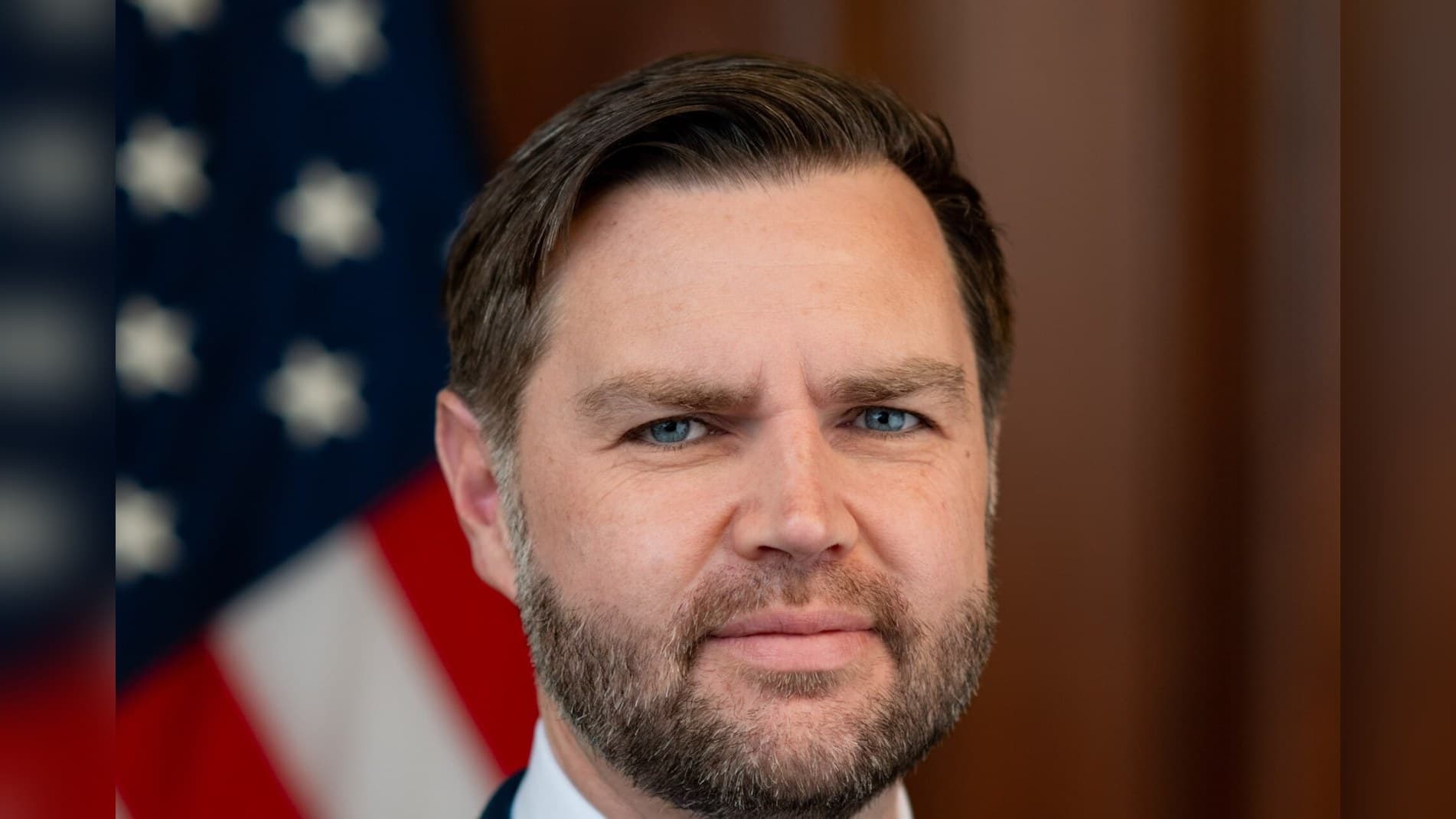 Official portrait of Vice President JD Vance in a dark suit in front of an American flag.