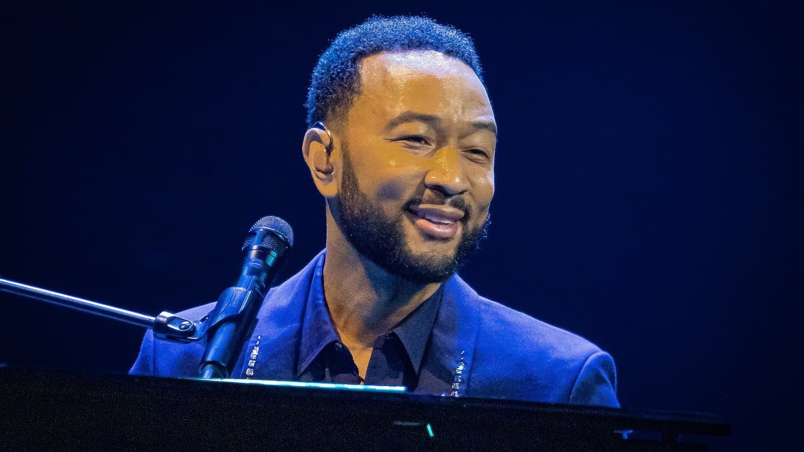 John Legend smiles while seated at a piano behind a microphone, wearing a dark blue jacket under deep blue stage lighting.