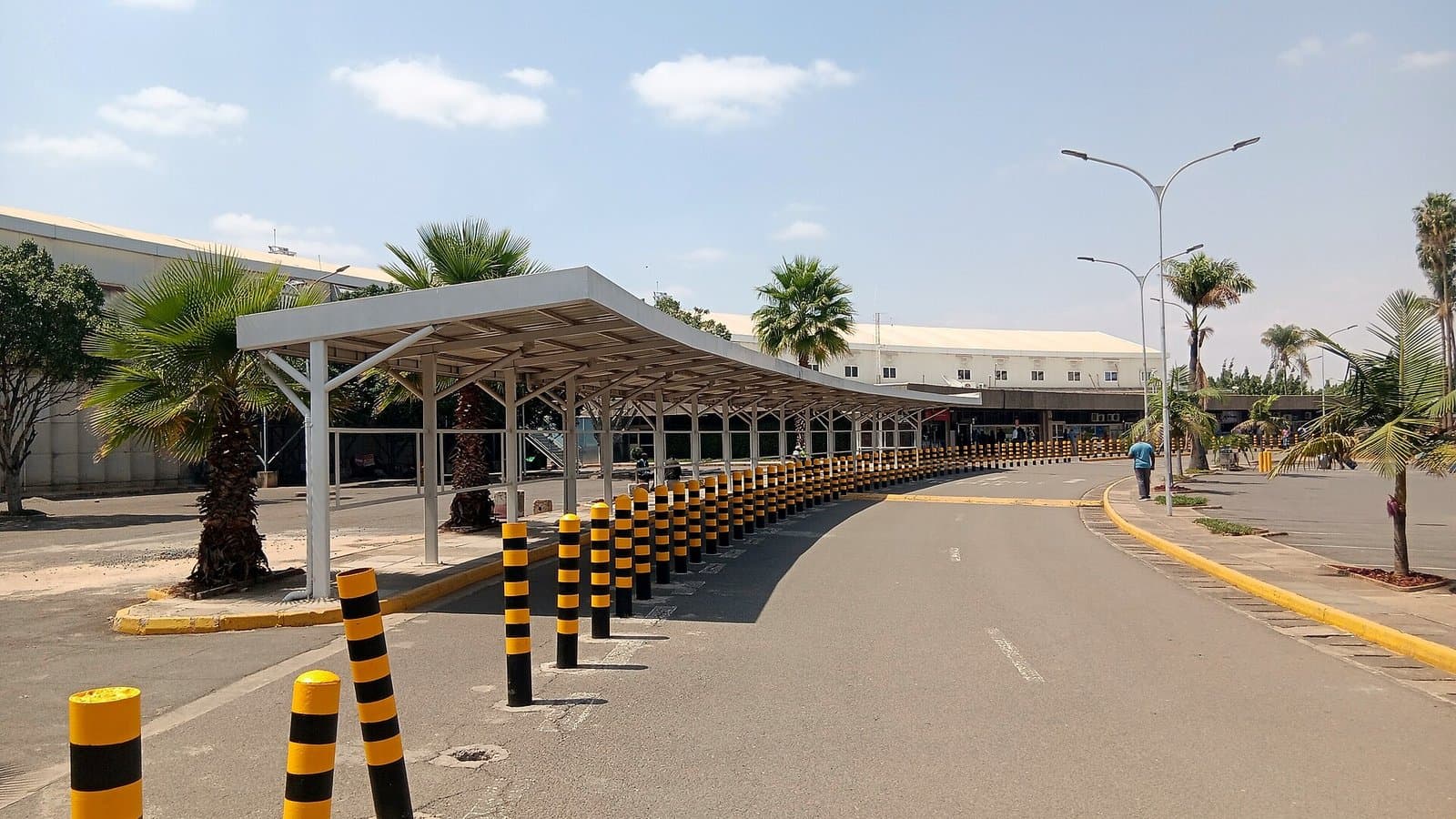 The landside approach to the terminal at Jomo Kenyatta International Airport in Nairobi, with a covered walkway, palm trees and yellow-and-black bollards.