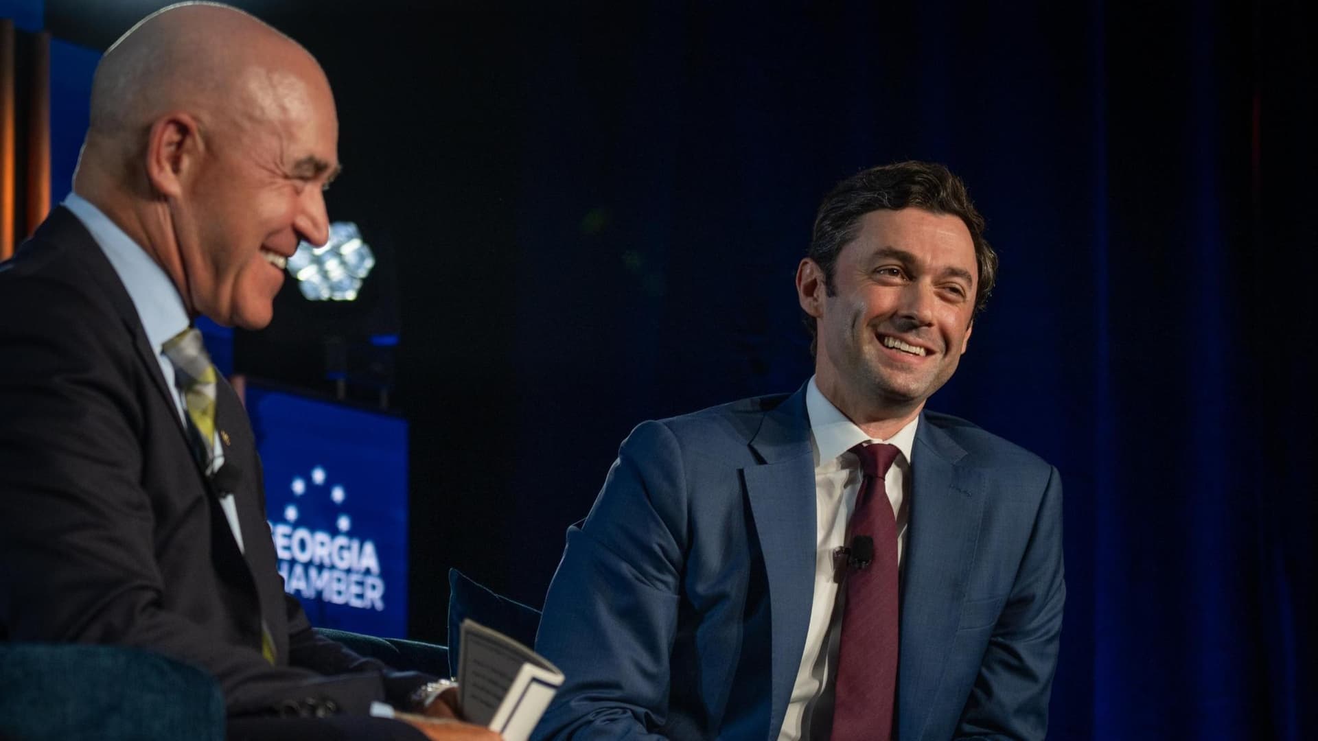 Senator Jon Ossoff in a blue suit and burgundy tie, smiling while seated on stage beside a moderator, with a Georgia Chamber sign in the background.