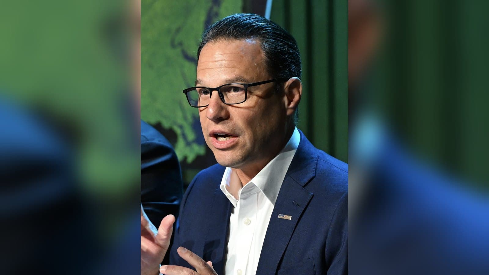 Pennsylvania Governor Josh Shapiro, wearing glasses and a dark suit with an open-collar white shirt, gestures as he speaks at a meeting.