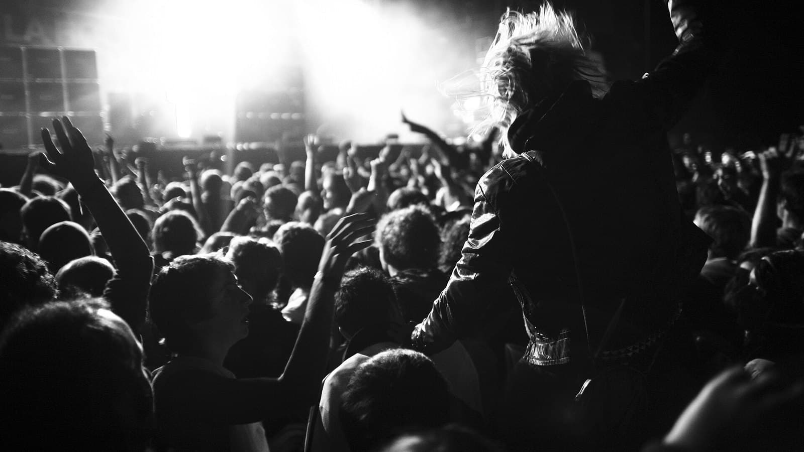 Black-and-white photo of a packed crowd with raised arms at a Justice concert, a bright stage behind them and a long-haired fan in the foreground.