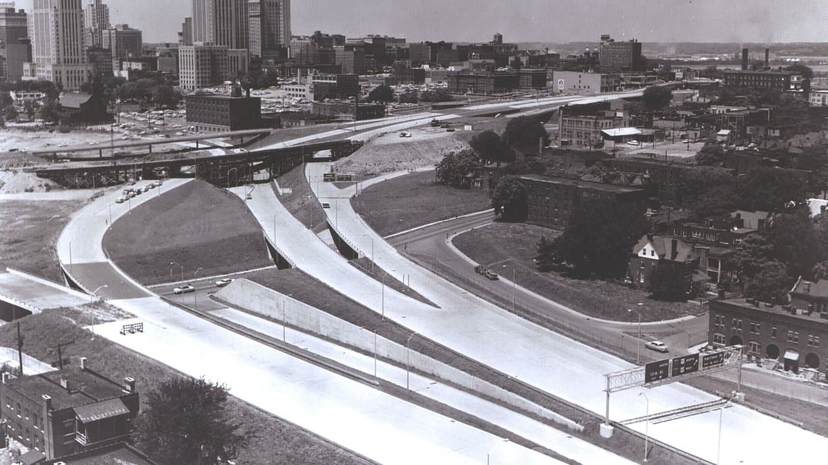Black-and-white aerial view of curving interstate ramps and overpasses leading into downtown Kansas City, with office towers in the distance.