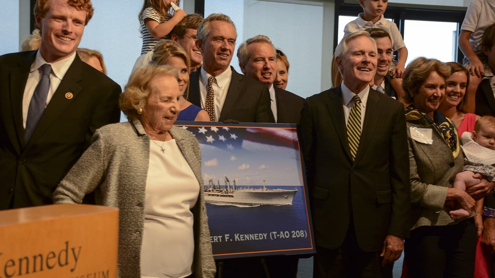 Ethel Kennedy, an elderly woman in a gray jacket, stands with smiling Kennedy family members and Navy Secretary Ray Mabus beside a framed picture of the ship USNS Robert F. Kennedy